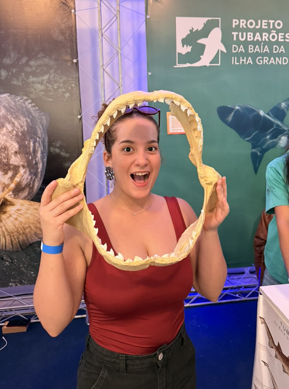Visitante segura a réplica impressa em 3D da mandíbula de tubarão-tigre diante do painel do Projeto Tubarões da Baía da Ilha Grande